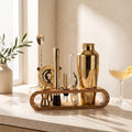 Cocktail-Set in Gold mit Shaker, Barsieb, Stössel und Barlöffel im Bambusständer auf heller Arbeitsfläche am Fenster, daneben ein Cocktailglas