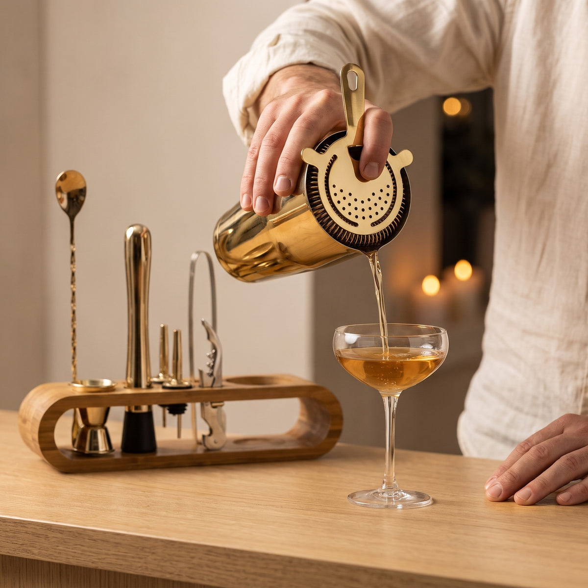 Hand giesst einen Drink aus dem goldenen Shaker durch das Barsieb in ein Cocktailglas, daneben das Set im Bambusständer